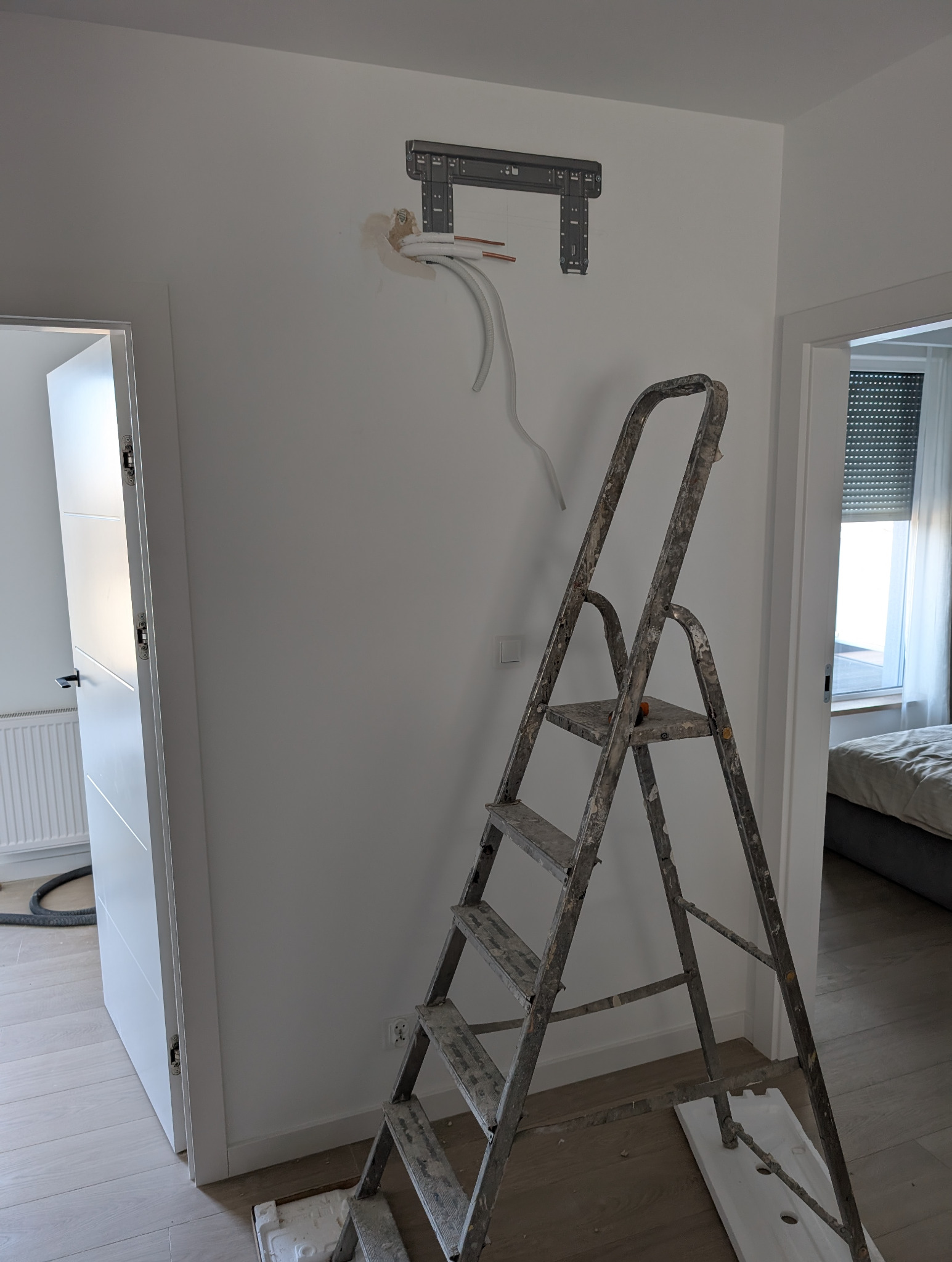 Montaż klimatyzacji: uchwyt na ścianie, rurki instalacyjne i drabina malarska w jasnym wnętrzu. Widoczne przygotowania do instalacji jednostki wewnętrznej.