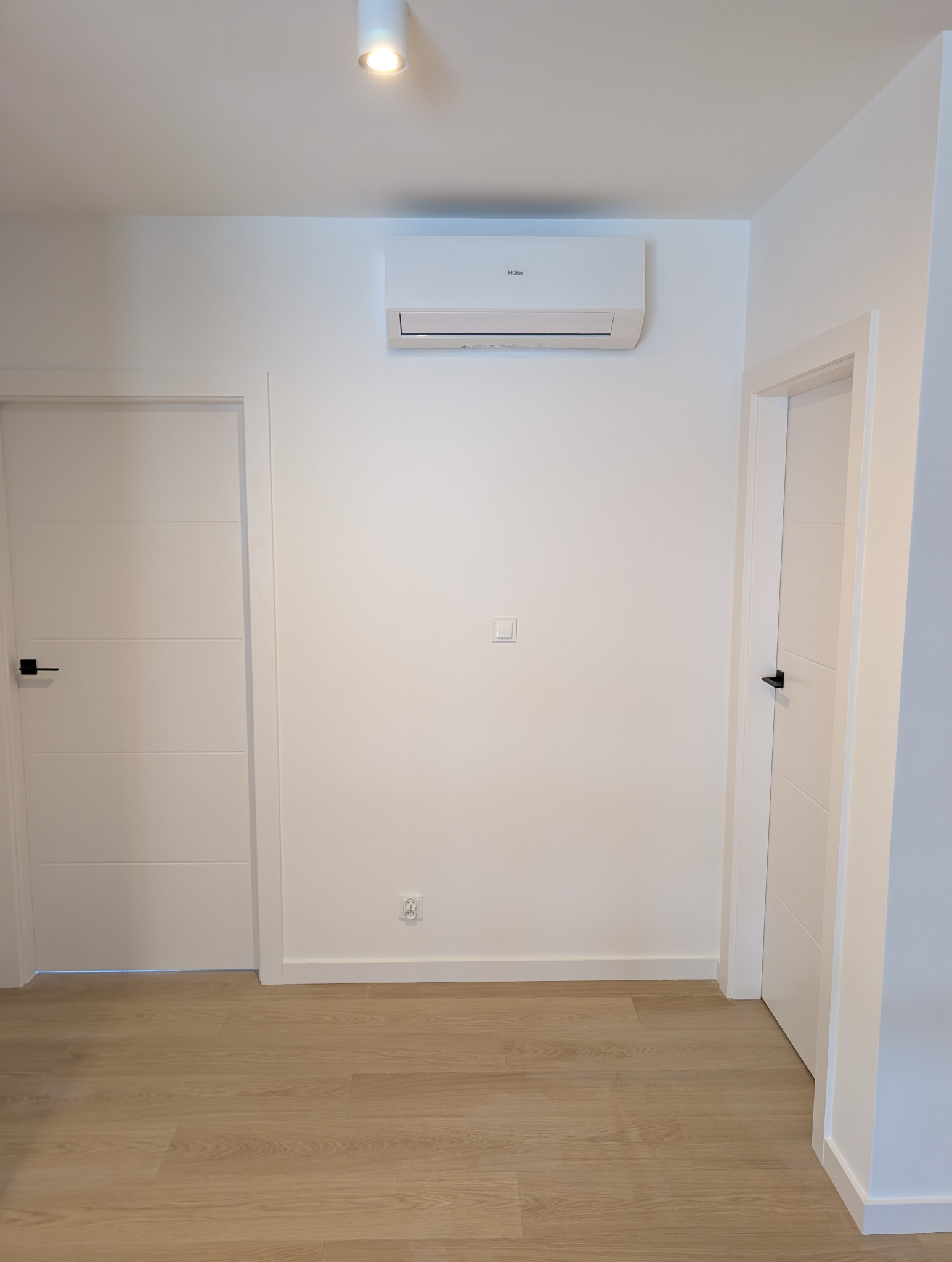 Biała klimatyzacja Haier zamontowana na ścianie w nowoczesnym wnętrzu z dwiema białymi drzwiami i drewnianą podłogą. Minimalistyczny design.