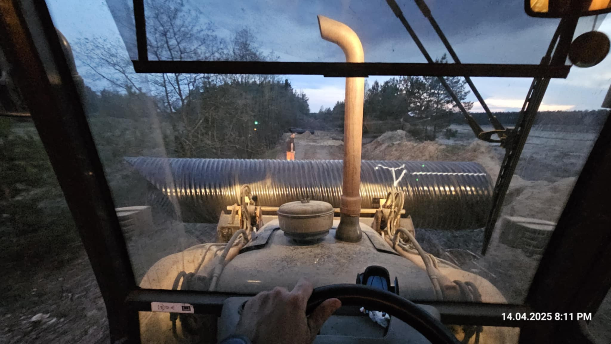 Widok z kabiny maszyny budowlanej na ułożony, długi drenaż rurowy w wykopie, z operatorem trzymającym kierownicę. W tle widoczny pracownik i usypana ziemia.