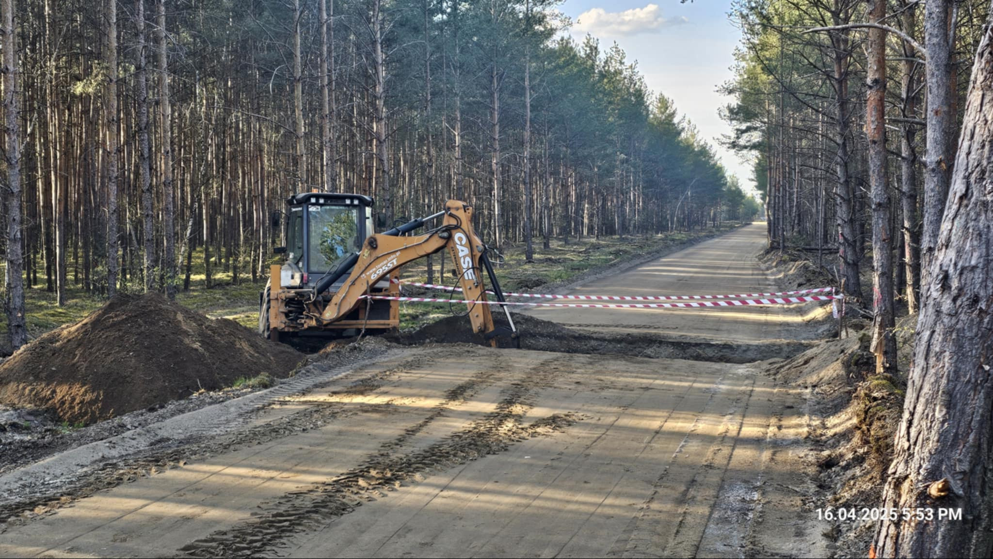 Żółta koparko-ładowarka CASE podczas wykopu w piaszczystym terenie leśnym. Wykop zabezpieczony taśmą ostrzegawczą, obok hałda ziemi. Widoczne ślady opon.