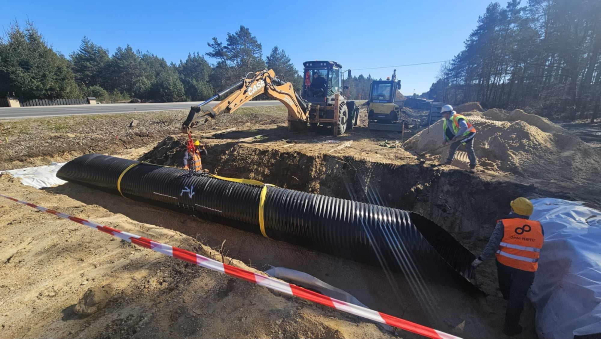 Układanie rury drenażowej w wykopie. Na budowie pracują dwie maszyny i dwóch robotników w kamizelkach odblaskowych. W tle drzewa i błękitne niebo.