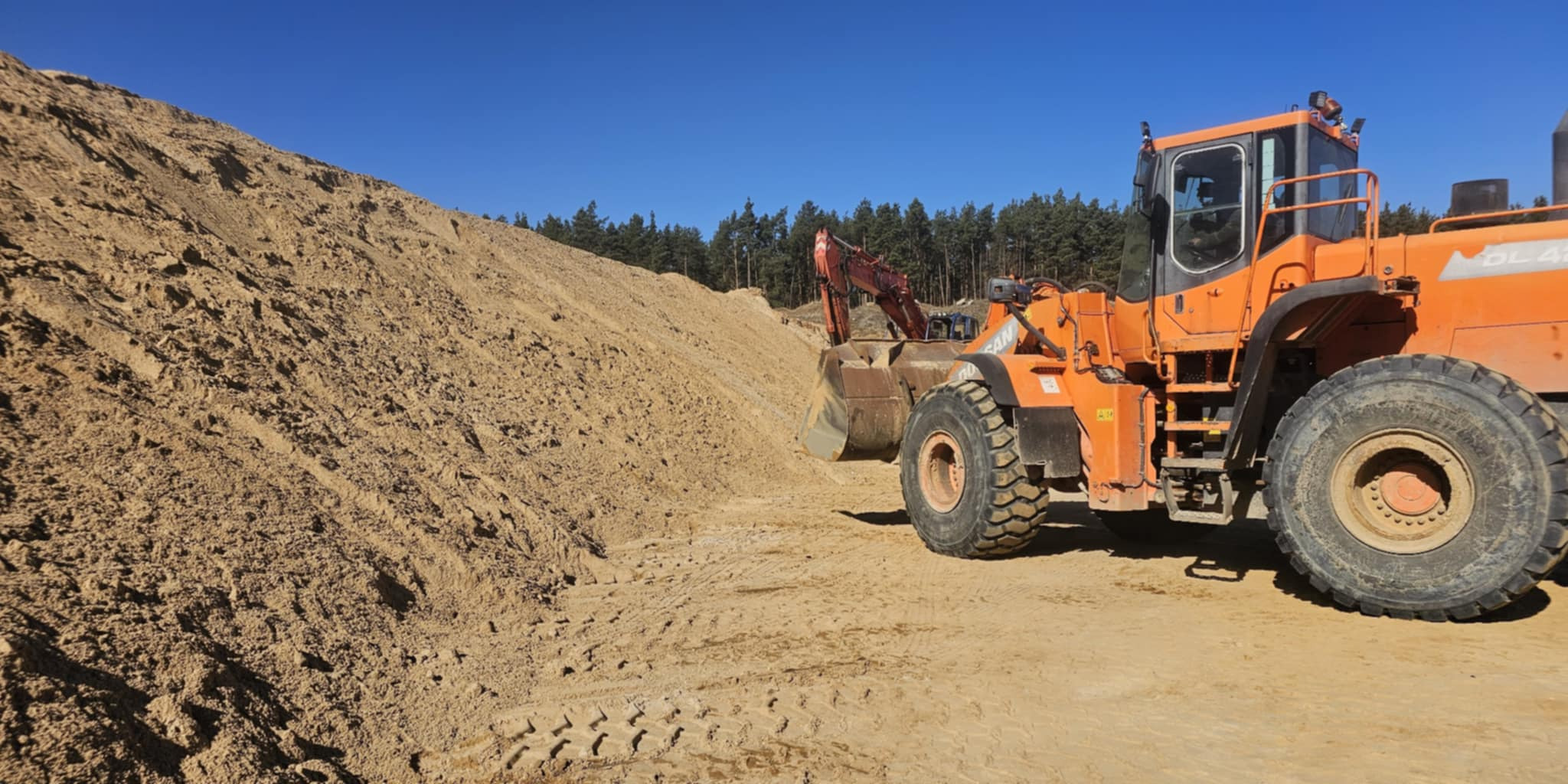 Żabia Wola: Pomarańczowa ładowarka kołowa Doosan przy hałdzie piasku. W tle widoczna koparka i las. Błękitne niebo.