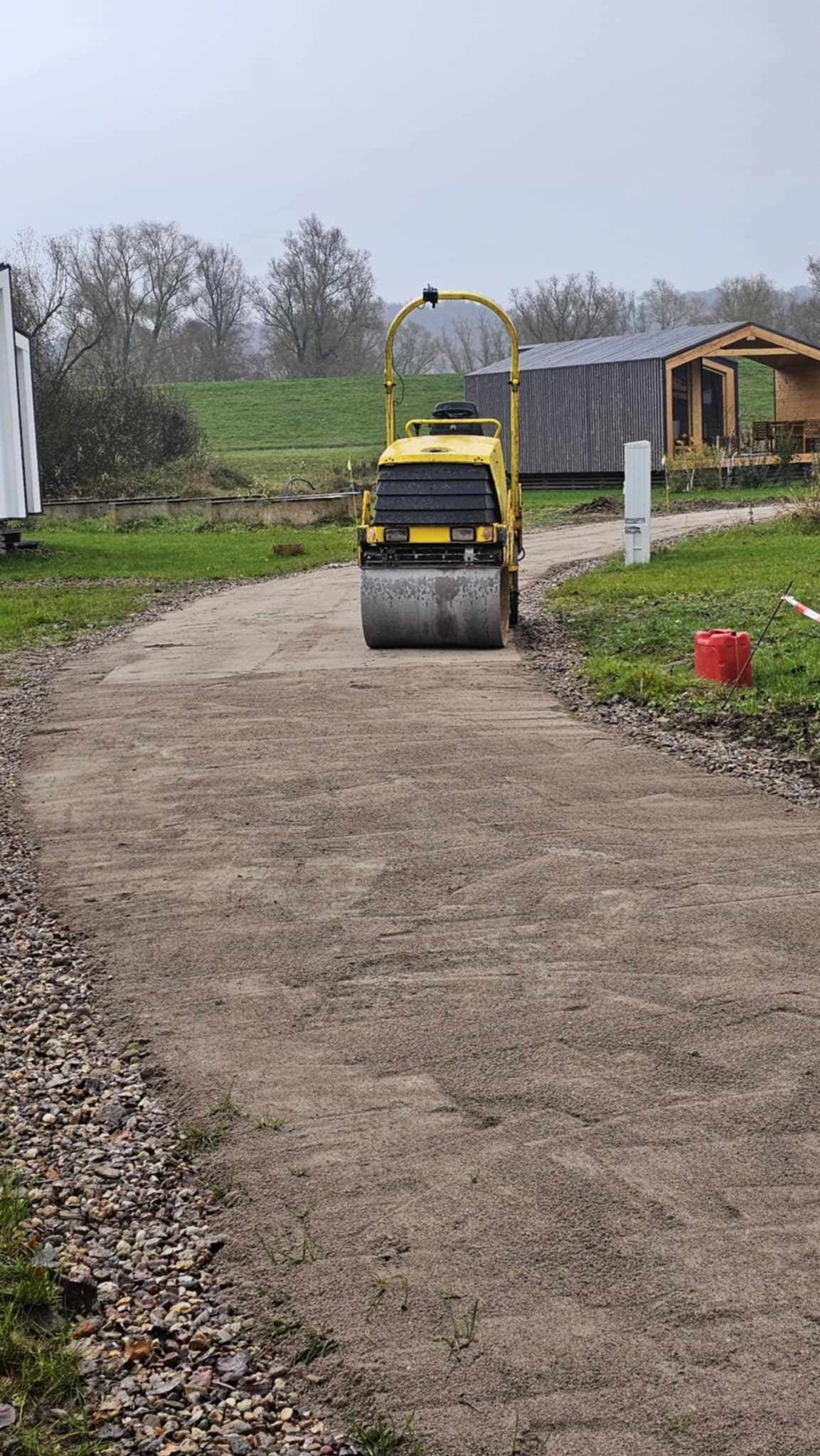 Żółty walec drogowy ubija piasek na ścieżce, w tle budynek z ciemnego drewna i zielone wzgórze. Praca ziemna z użyciem ciężkiego sprzętu.