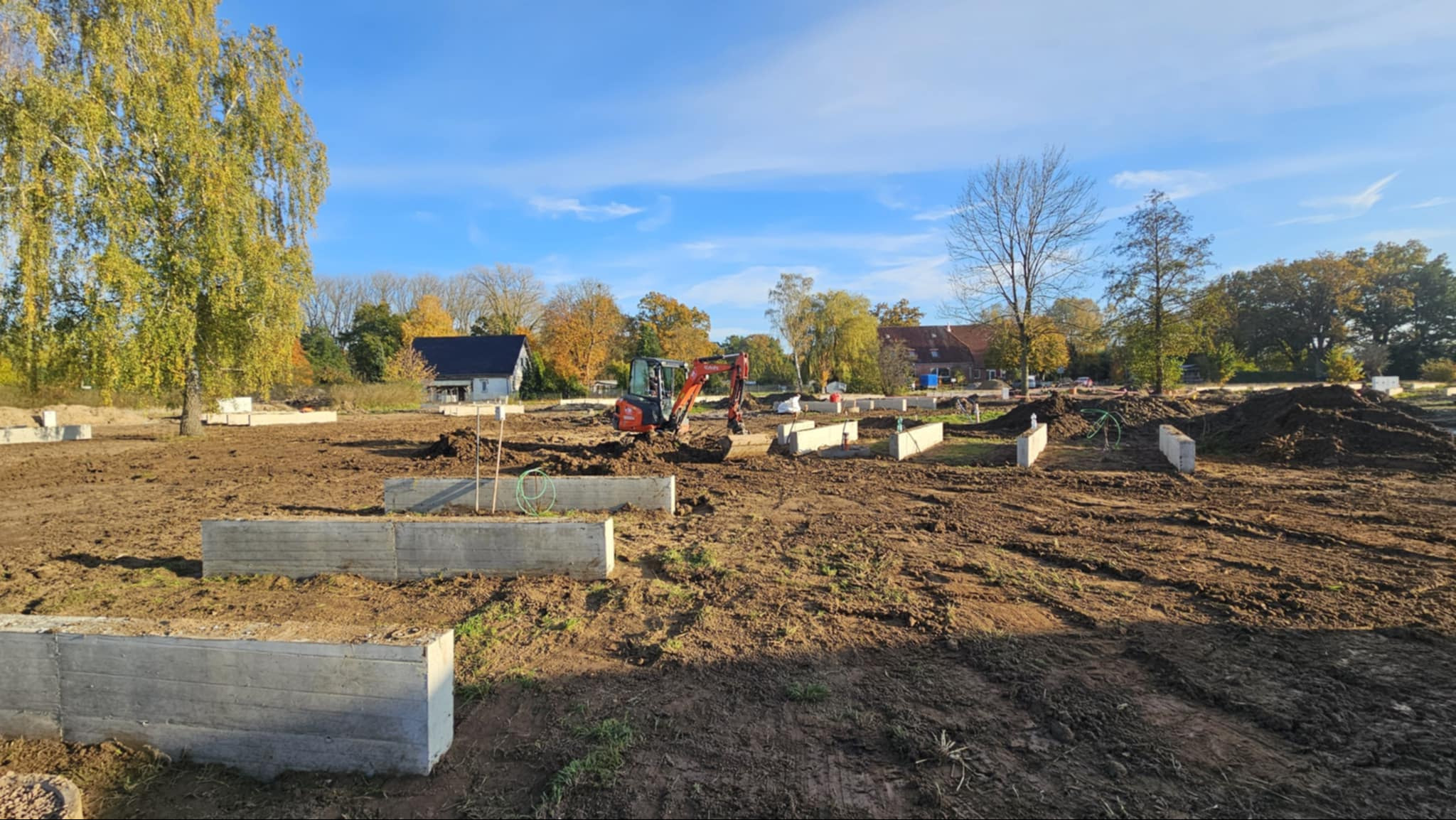 Fundamenty pod budowę domu jednorodzinnego z użyciem mini koparki. Widoczne betonowe szalunki i przygotowany teren. Jesienna sceneria, błękitne niebo.