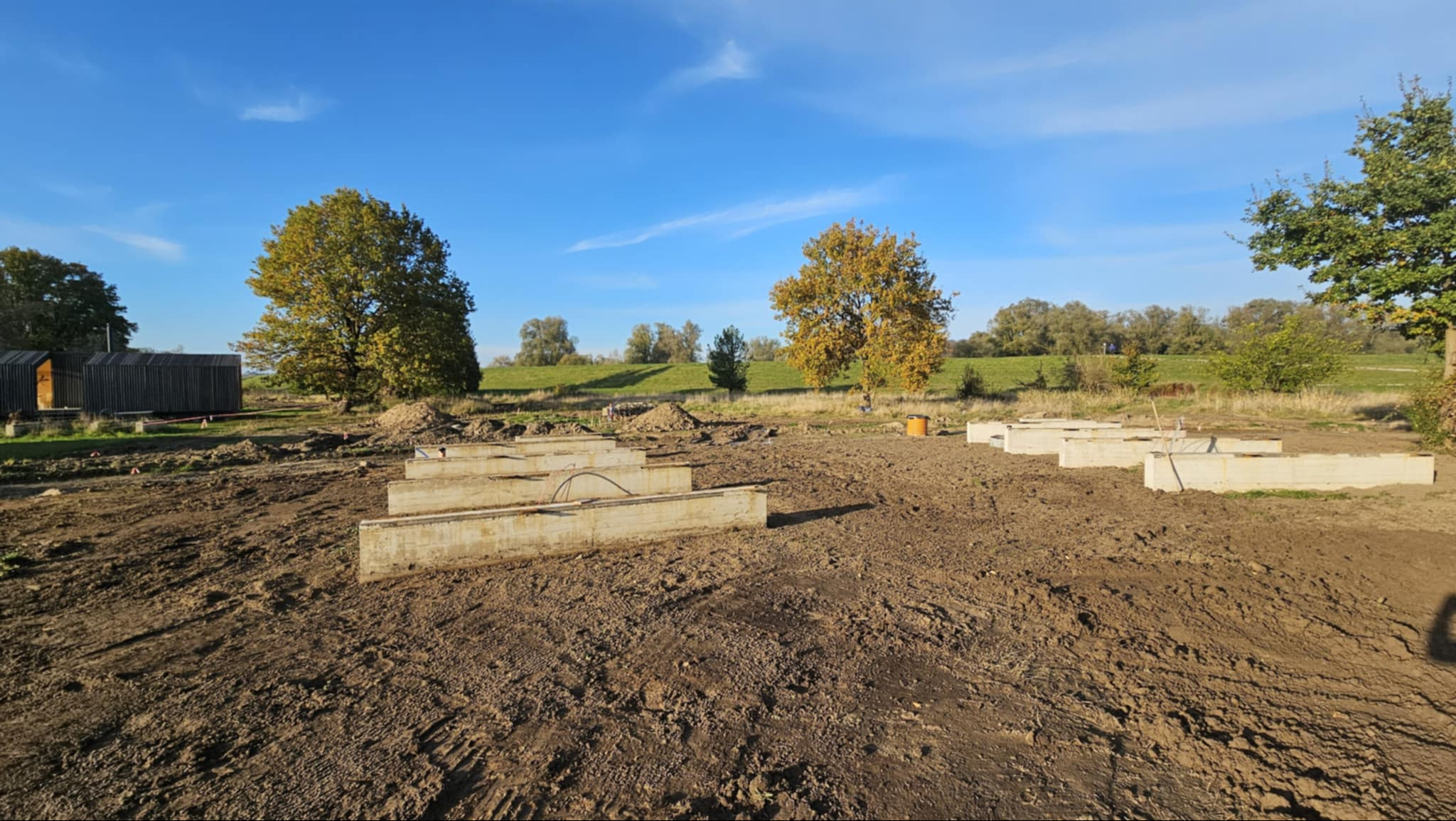 Betonowe ławy fundamentowe na działce z widokiem na drzewa i błękitne niebo. Widoczne przewody w fundamencie. Prace ziemne w toku, teren przygotowany pod budowę.