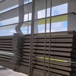 Zamknięte białe rolety poziome z widokiem na zewnątrz. Szare lamele, sznurki do regulacji i taśma podtrzymująca. Widok na drzewa i niebo przez szczeliny.