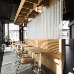Jasne, zwiewne zasłony panelowe dzielące przestrzeń restauracji. Naturalne światło podkreśla minimalistyczny design z drewnianymi elementami i metalowymi detalami.
