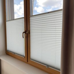 Home and Industry Automation Kamil Korziuk - Okno z drewnianą ramą i zamontowanymi plisami w kolorze białym. Z boku widoczne fragmenty zasłon w odcieniach beżu. Widok na błękitne niebo.