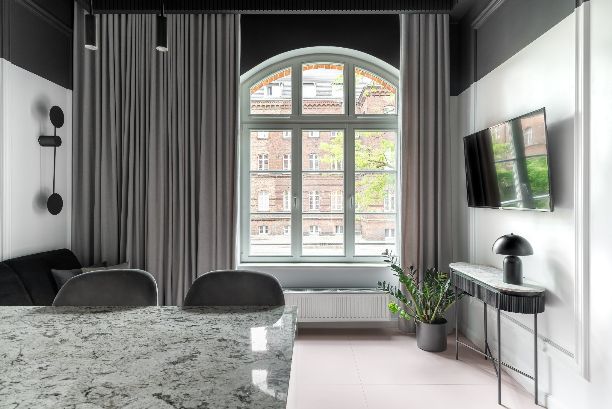 Eleganckie, szare zasłony w nowoczesnym wnętrzu z oknem w kształcie łuku, przez które widać ceglany budynek. Minimalistyczny wystrój z czarnymi akcentami i marmurowym stołem.