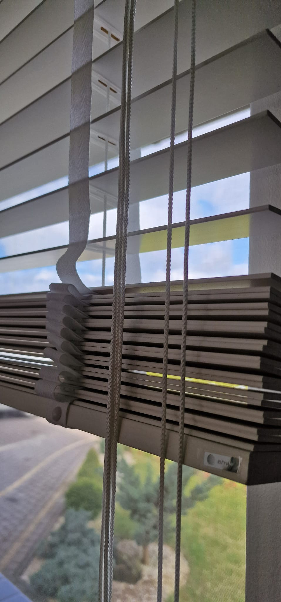 Zamknięte białe rolety poziome z widokiem na zewnątrz. Szare lamele, sznurki do regulacji i taśma podtrzymująca. Widok na drzewa i niebo przez szczeliny.