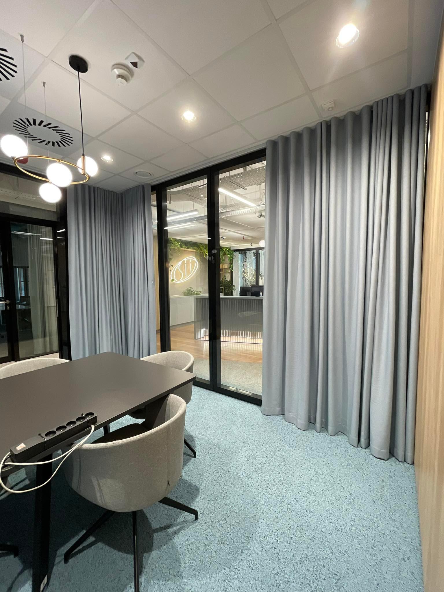 Jasnoszare, długie zasłony w nowoczesnym biurze z przeszklonymi ścianami, biurkiem i krzesłami. Widoczne oświetlenie sufitowe.