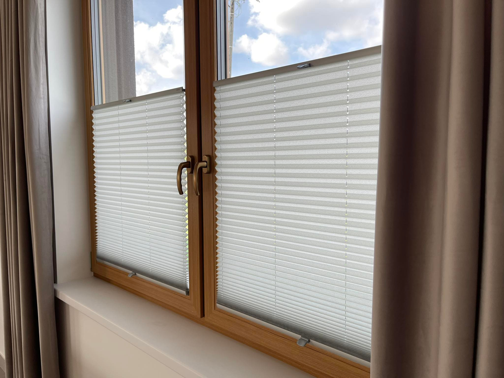 Okno z drewnianą ramą i zamontowanymi plisami w kolorze białym. Z boku widoczne fragmenty zasłon w odcieniach beżu. Widok na błękitne niebo.