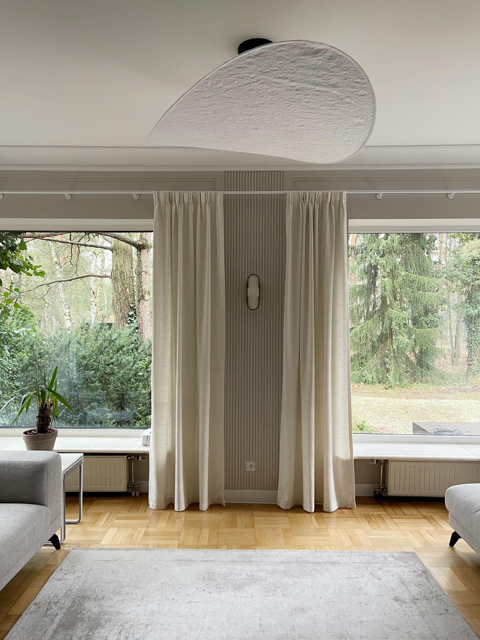 Eleganckie, kremowe zasłony w salonie z parkietem i dywanem. Duże okno z widokiem na zieleń. Nowoczesna lampa sufitowa i stonowana kolorystyka.