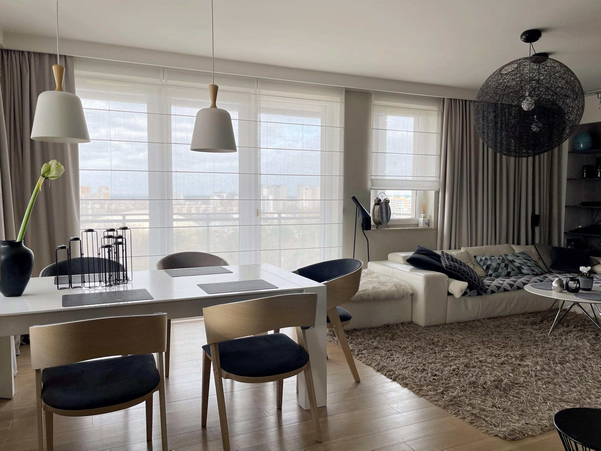 Jasne wnętrze salonu z widokiem na miasto przez okna przesłonięte jasnymi roletami rzymskimi. Minimalistyczny stół i krzesła na pierwszym planie.