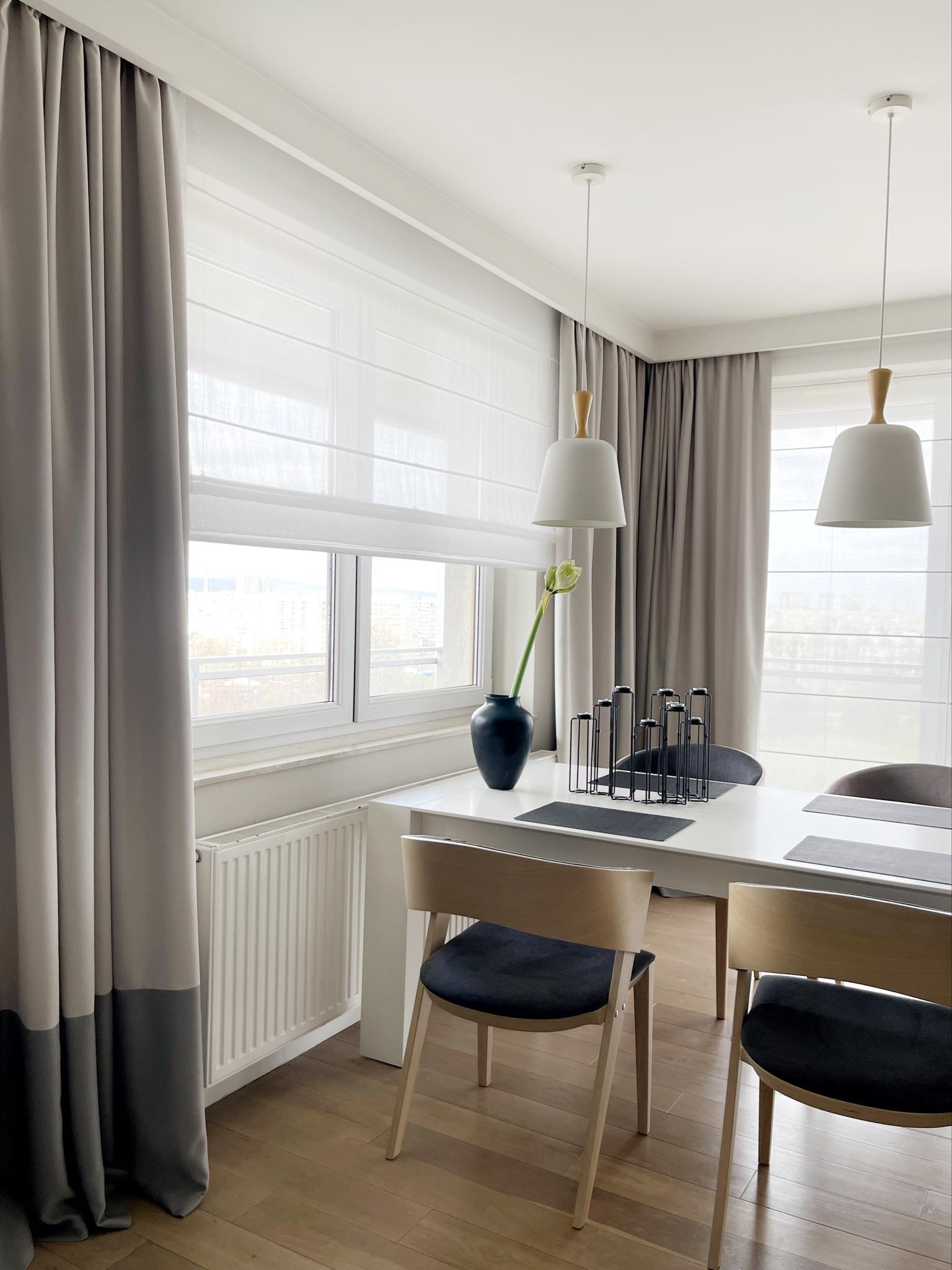 Eleganckie wnętrze: szare zasłony z pasem, rolety rzymskie w oknie, biały stół z dekoracjami. Stonowana kolorystyka i minimalistyczny design.