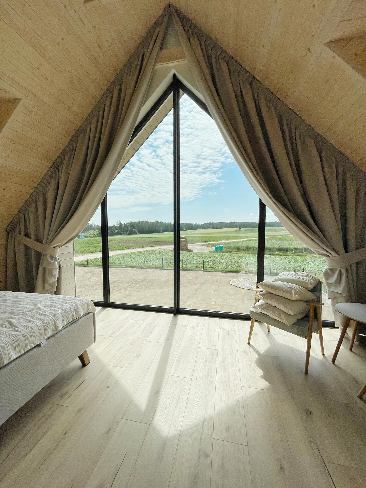 Beżowe zasłony w trójkątnym oknie na poddaszu z widokiem na zielone pole. Naturalne światło słoneczne pada na podłogę. Wnętrze w stylu skandynawskim.
