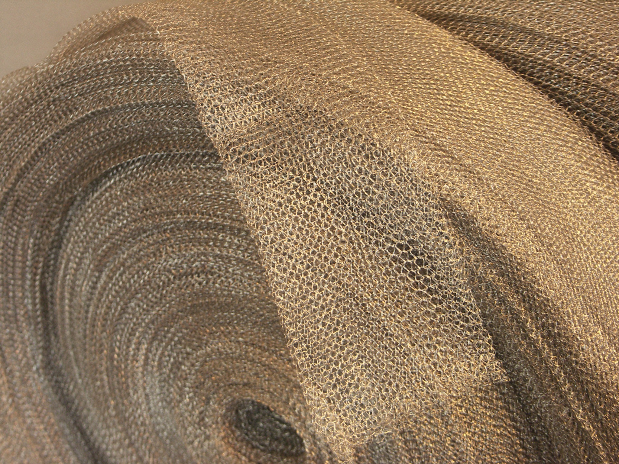 Zwinięta, metalowa siatka filtracyjna o drobnych oczkach, w odcieniach szarości i złota, układająca się warstwami. Tekstura siatki jest wyraźnie widoczna.