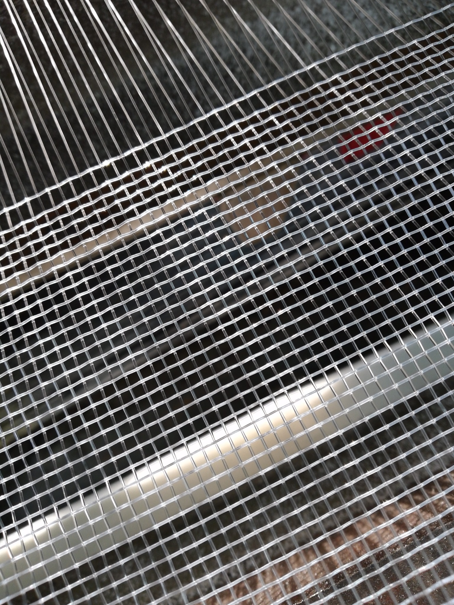 Siatka filtracyjna o drobnych oczkach, ukośnie ułożona, z widocznymi cienkimi, metalowymi prętami w tle. Element studni głębinowej.