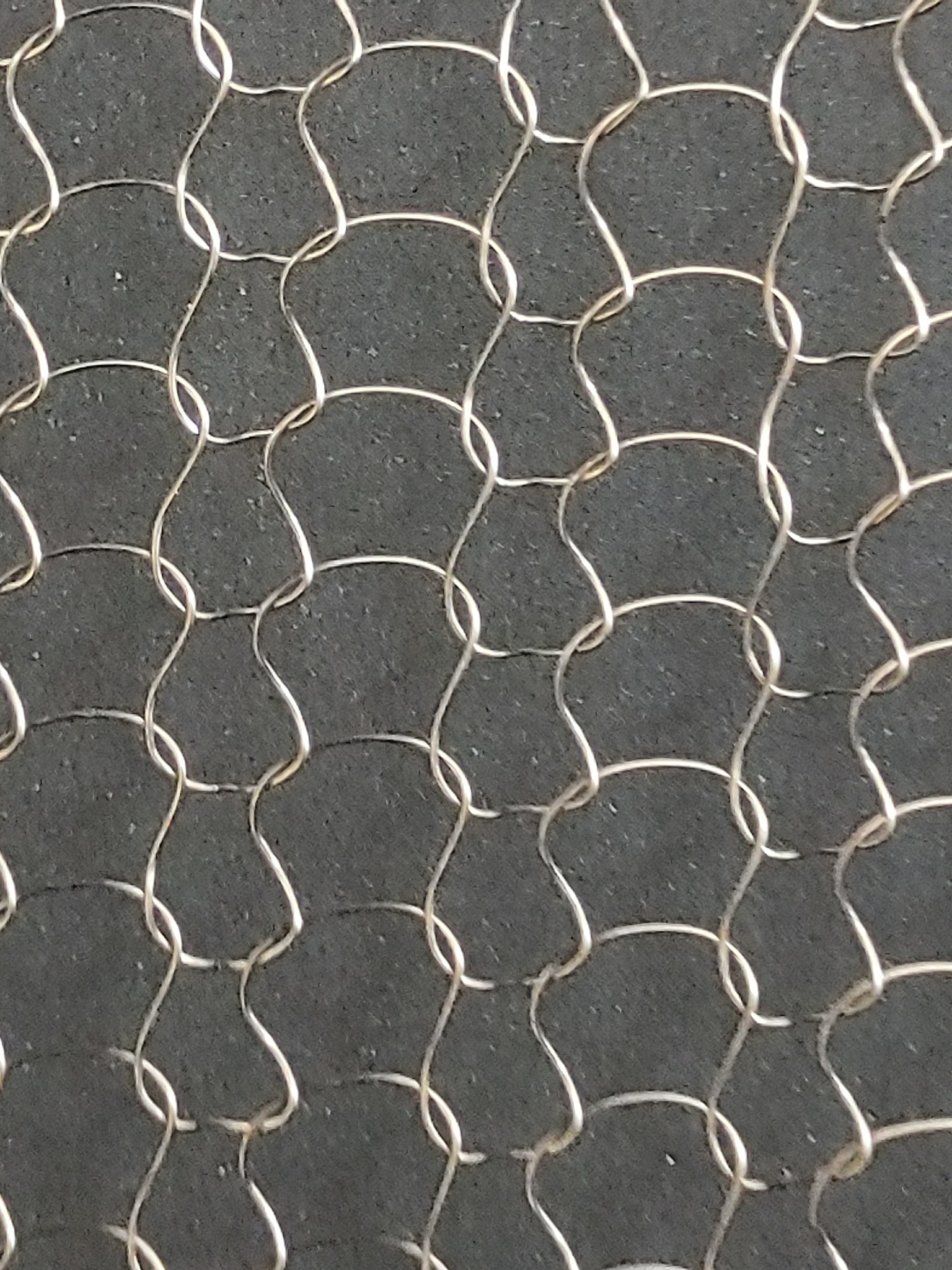 Metalowa siatka o nieregularnych oczkach na tle szarej, chropowatej powierzchni. Detal z bliska, ukazujący strukturę materiału.