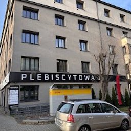 UnicSystem Centrum Pomocy Psychologicznej Dawid Stasiak - Elewacja budynku z szyldem 'Plebiscytowa 1' i żółtą budką. Widok na fasadę w słoneczny dzień, z zaparkowanymi autami i fragmentami drzew.