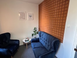 Przytulny gabinet z aksamitną sofą w odcieniach granatu, stolikiem kawowym i rośliną doniczkową. Na ścianie grafiki w białych ramkach i panel dekoracyjny.