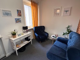 Przytulny gabinet: białe ściany, niebieski fotel i sofa, biurko z lampką i rośliną. Okno z żółtą zasłoną. Dekoracje ścienne. Idealne miejsce na spotkanie.