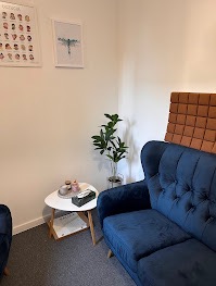 Przytulny gabinet z niebieską sofą, stolikiem i rośliną. Na ścianie grafika z wizerunkami twarzy i obrazek z ważką. Czyste, jasne wnętrze.