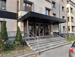 Wejście do prywatnej kliniki z widocznymi schodami, poręczami i tablicą informacyjną. Budynek z szarej cegły, dzień, widoczne krzewy i fragment samochodu.