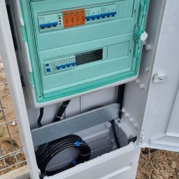 FEMAVOLT - Zewnętrzna, otwarta biała skrzynka elektryczna z zieloną osłoną modułów, pomarańczowymi ogranicznikami przepięć SIMTEC i schludnie ułożonymi kablami zasilającymi na ziemi.