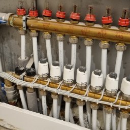 FEMAVOLT - Mosiężny rozdzielacz ogrzewania podłogowego MAINCOR z białymi zaworami i przepływomierzami, obok otwartej puszki z kolorowymi przewodami elektrycznymi.