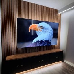 FEMAVOLT - Nowoczesna ściana RTV z pionowymi drewnianymi lamelami, dużym telewizorem i czarną szafką z podświetleniem LED, harmonijnie wkomponowana w minimalistyczne wnętrze.