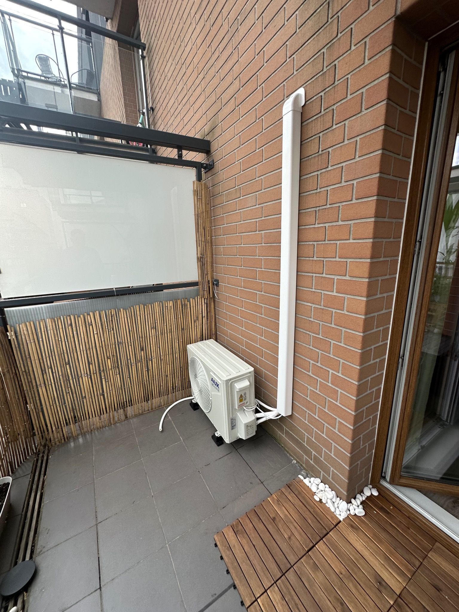 Klimatyzator AUX zamontowany na balkonie z ceglaną ścianą, z widoczną białą rurą odprowadzającą. Podłoże wyłożone płytkami i drewnianymi panelami.