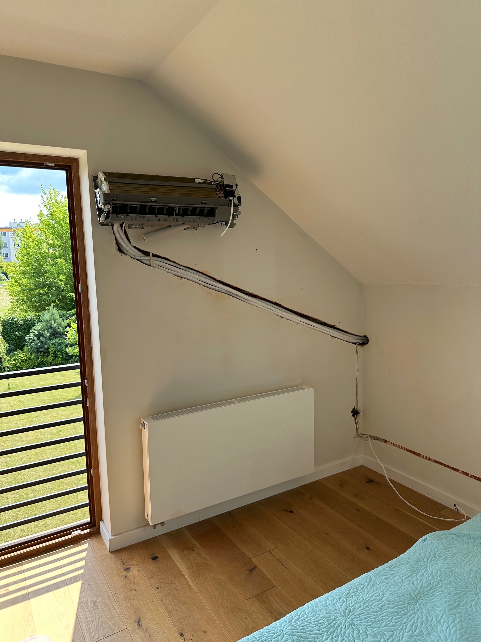 Montaż klimatyzacji w pokoju na poddaszu. Widoczne przewody prowadzone w ścianie, okno balkonowe i grzejnik. Częściowo zdemontowana obudowa klimatyzatora.
