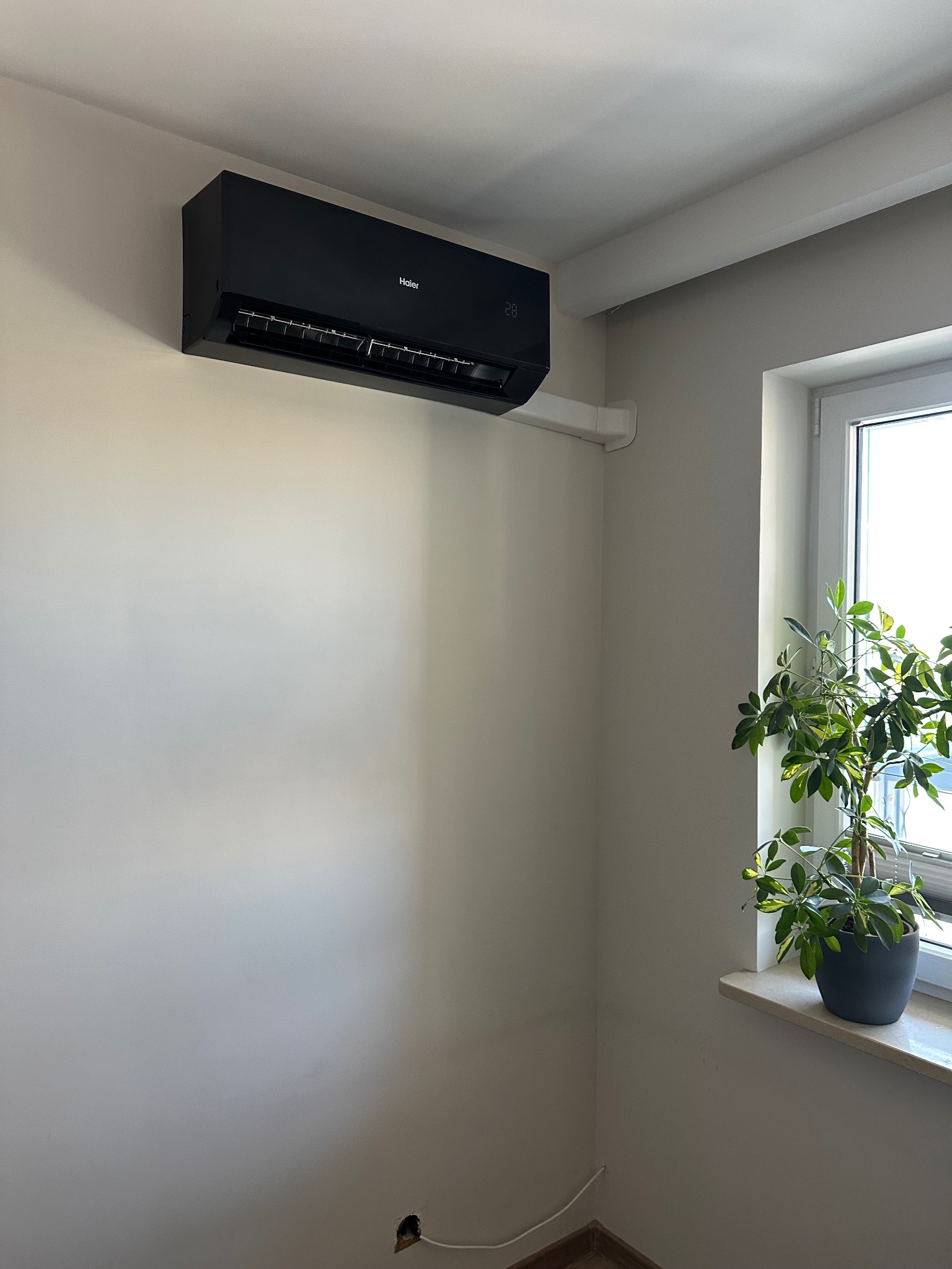 Czarny klimatyzator Haier zamontowany na ścianie w pokoju, obok okna z rośliną w doniczce. Widoczne białe rury instalacyjne i gniazdko elektryczne.