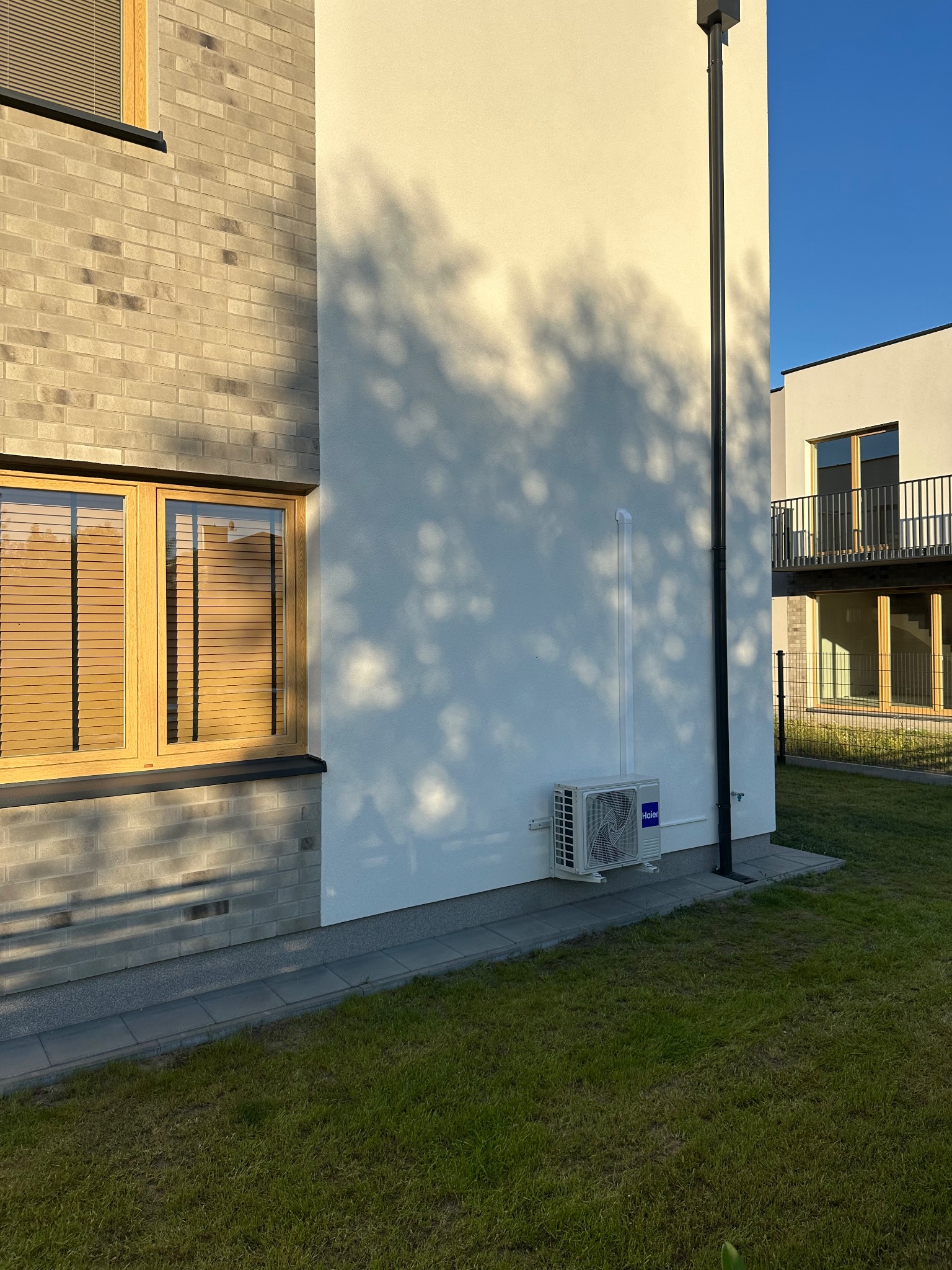 Biała jednostka klimatyzacji Haier zamontowana na elewacji budynku. Cień drzewa na ścianie. Widoczny fragment trawnika i budynku sąsiedniego.