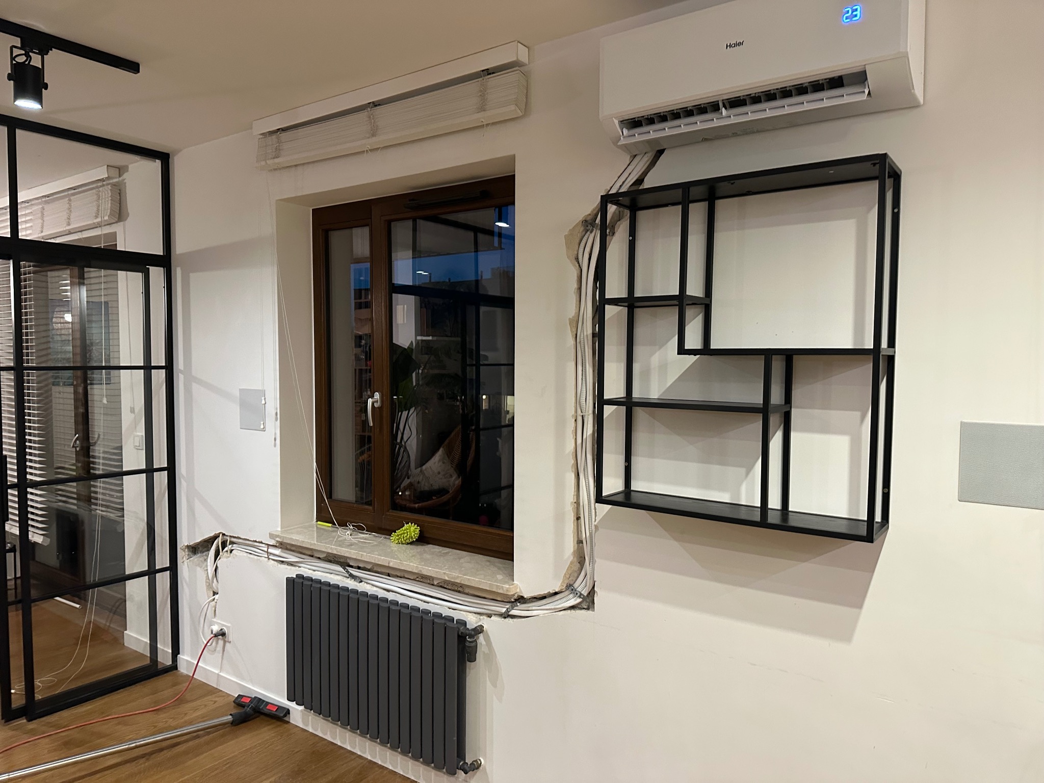 Montaż klimatyzacji w mieszkaniu: widoczne przewody i instalacja po montażu jednostki wewnętrznej, obok okna i grzejnika. Na ścianie półka dekoracyjna.