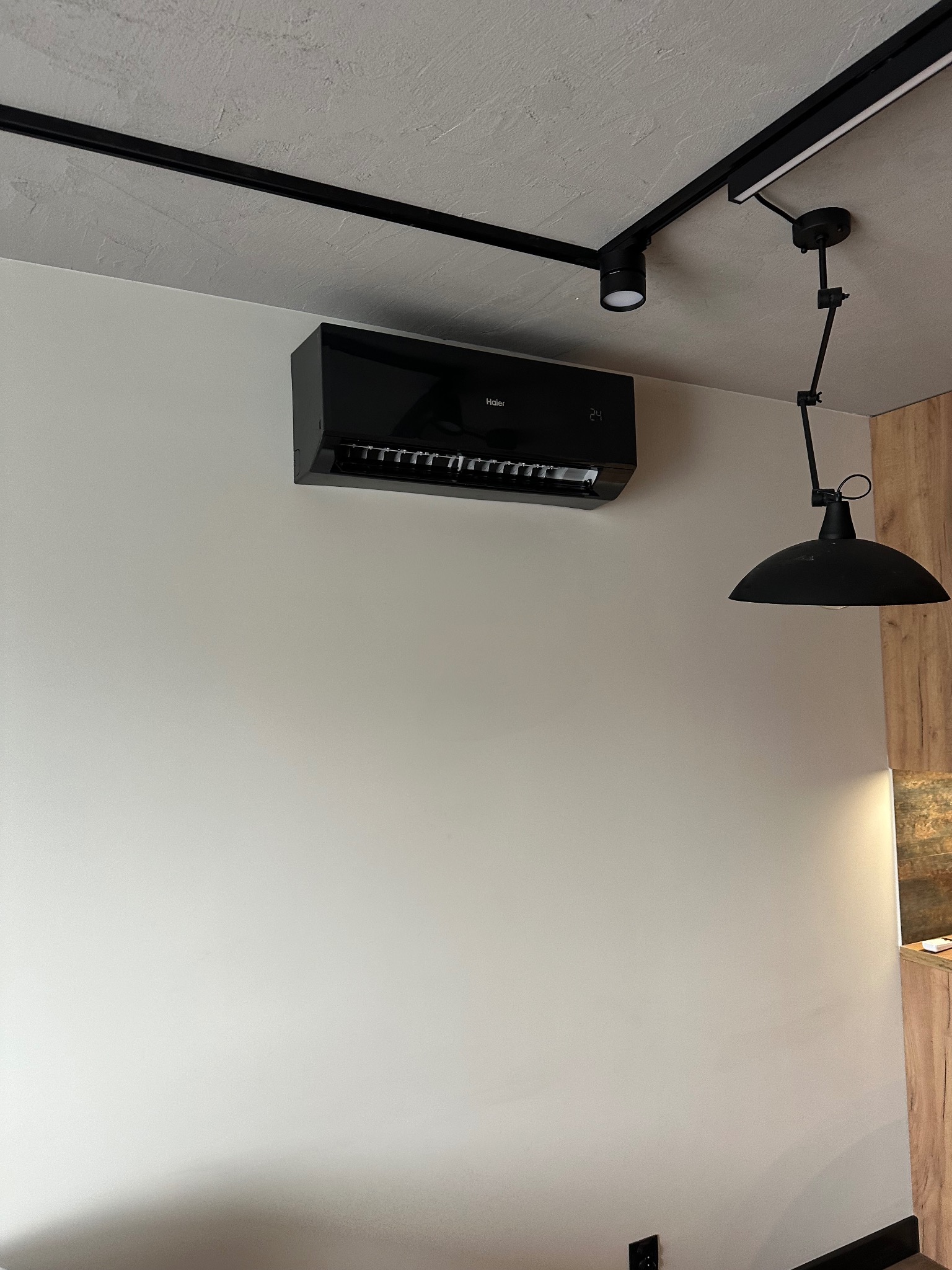 Czarny klimatyzator Haier na białej ścianie, podświetlony reflektorem na szynie i lampą wiszącą. Widoczny fragment drewnianej zabudowy.