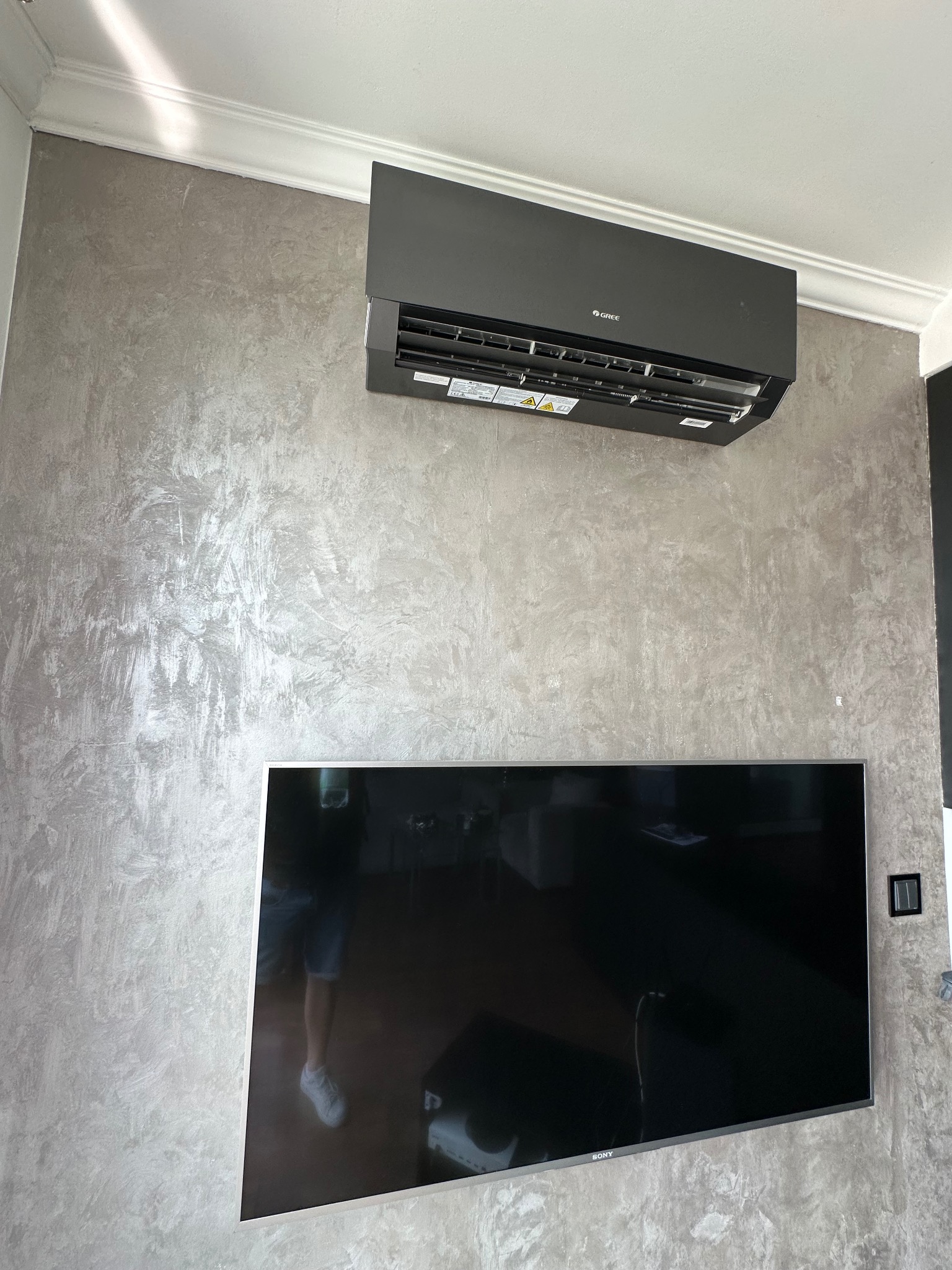 Czarny klimatyzator marki Gree zamontowany na ścianie o dekoracyjnej fakturze, poniżej telewizor Sony. Widoczny fragment sufitu z listwą dekoracyjną.