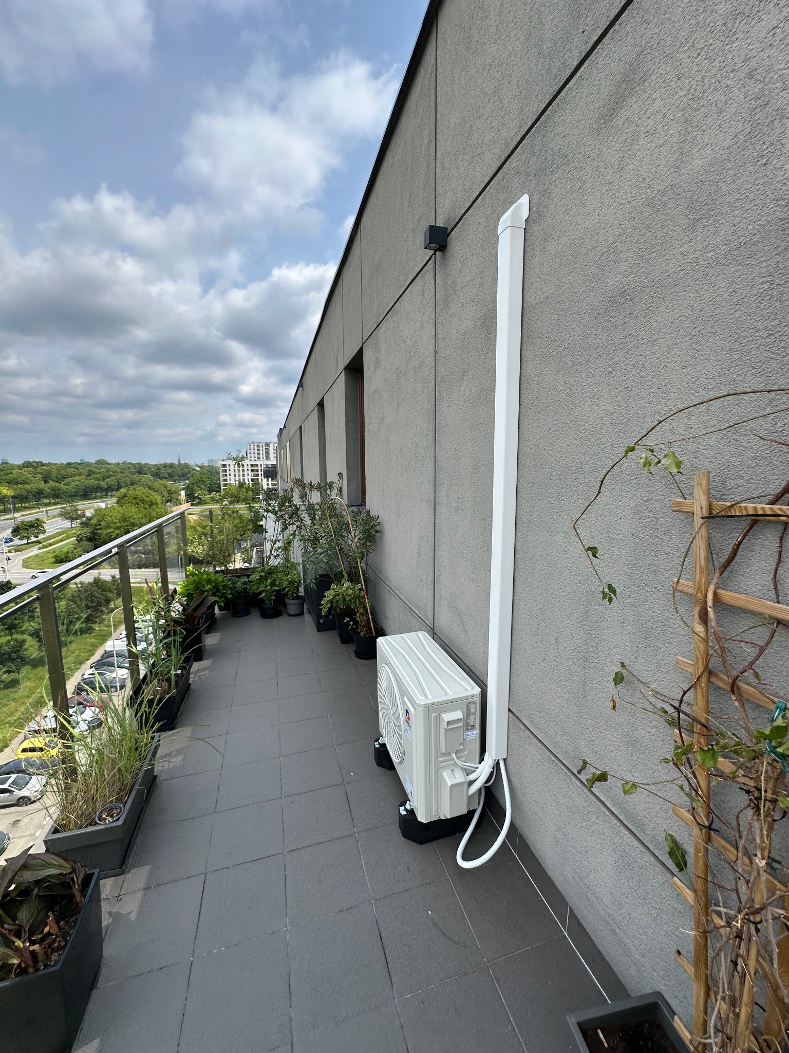 Klimatyzator zewnętrzny zamontowany na balkonie w Warszawie. Widoczna instalacja odprowadzająca skropliny. Balkon z roślinami w donicach i widok na miasto.