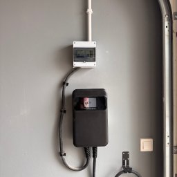 EL-Ryś - Zamontowana stacja ładowania EV z kablem, skrzynką elektryczną i peszlem na szarej ścianie garażu. Widoczny włącznik światła i metalowa rama bramy garażowej.