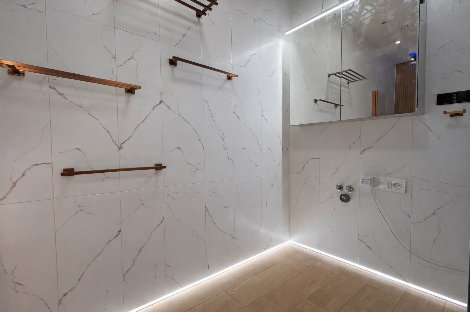 Nowoczesna łazienka z białymi płytkami imitującymi marmur, oświetleniem LED przy podłodze i miedzianymi relingami na ręczniki. Widoczne gniazdka i lustro z szafką.