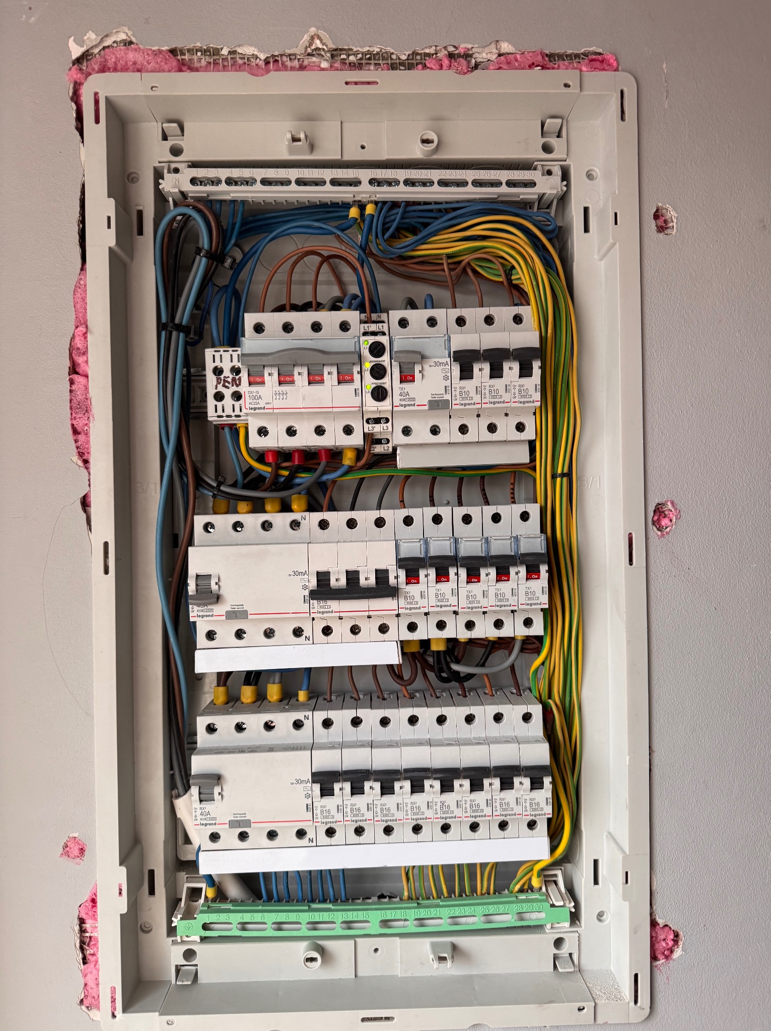Rozdzielnica elektryczna Legrand w trakcie montażu, widoczne kable, bezpieczniki i oznaczenia. Ściana z widocznymi śladami po kuciu i różową pianką izolacyjną.