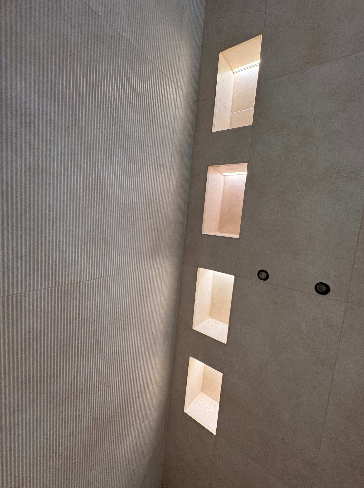 Ściana z beżowych płytek z czterema wnękami oświetlonymi od wewnątrz, oraz trzema okrągłymi punktami oświetleniowymi. Pionowe ułożenie płytek z delikatnym wzorem.