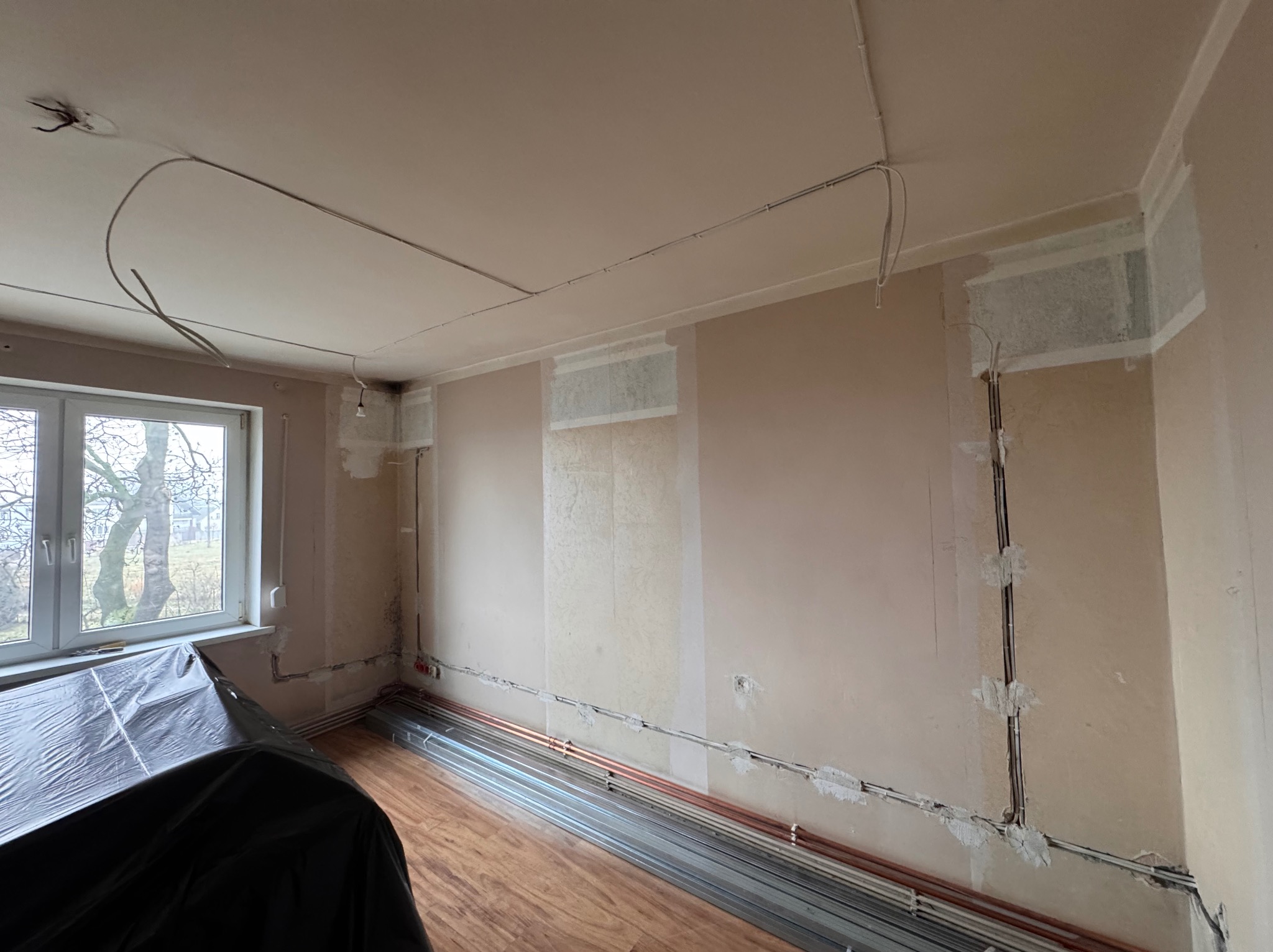 Wnętrze w trakcie remontu: widoczne przewody elektryczne na ścianach i suficie, rury grzewcze ułożone wzdłuż ściany, zabezpieczone meble folią. Etap prac instalacyjnych.