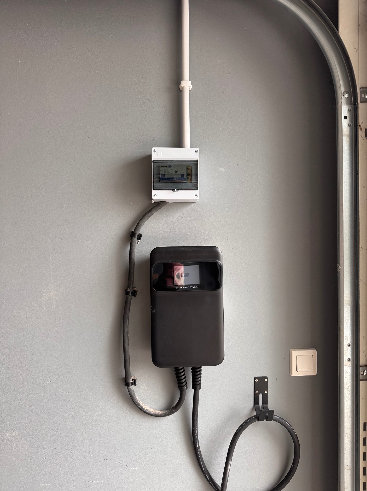 Zamontowana stacja ładowania EV z kablem, skrzynką elektryczną i peszlem na szarej ścianie garażu. Widoczny włącznik światła i metalowa rama bramy garażowej.