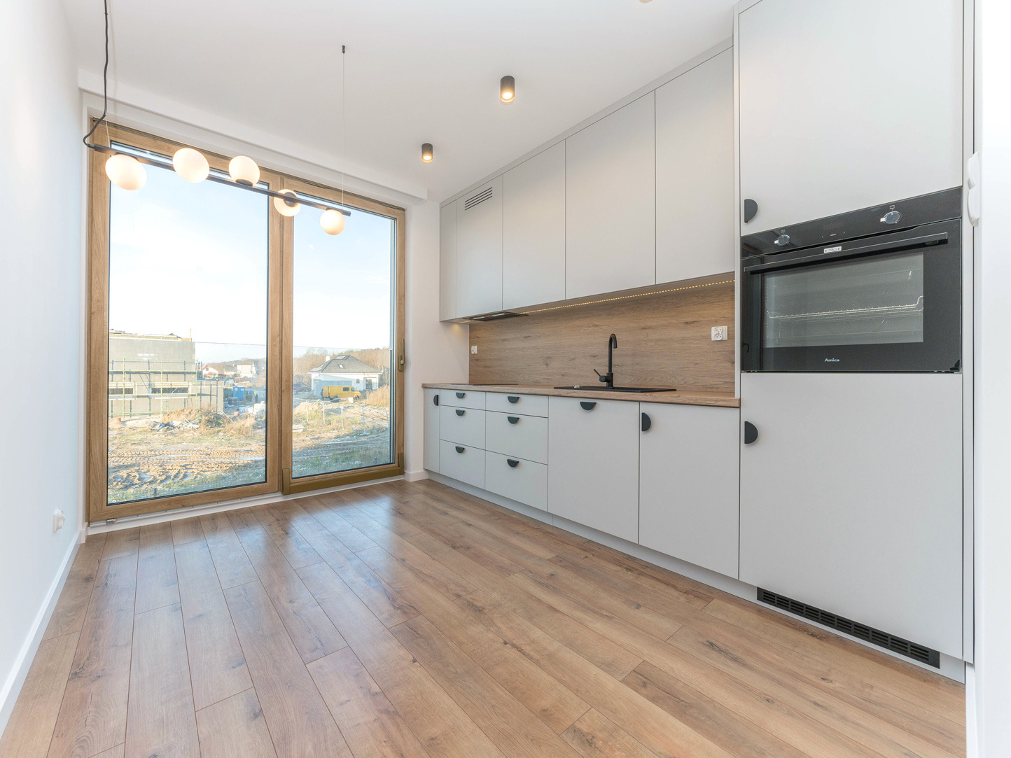 Jasna kuchnia z drewnianym blatem i dużym oknem z widokiem na plac budowy. Minimalistyczne szafki, drewniana podłoga i nowoczesne oświetlenie.