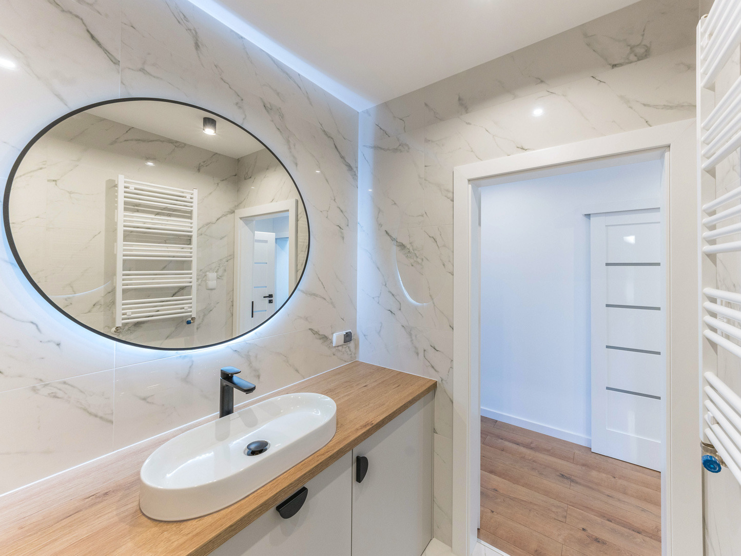 Nowoczesna łazienka z owalnym lustrem podświetlanym LED, marmurowymi płytkami i drewnianym blatem. Widoczna umywalka nablatowa i grzejnik drabinkowy.
