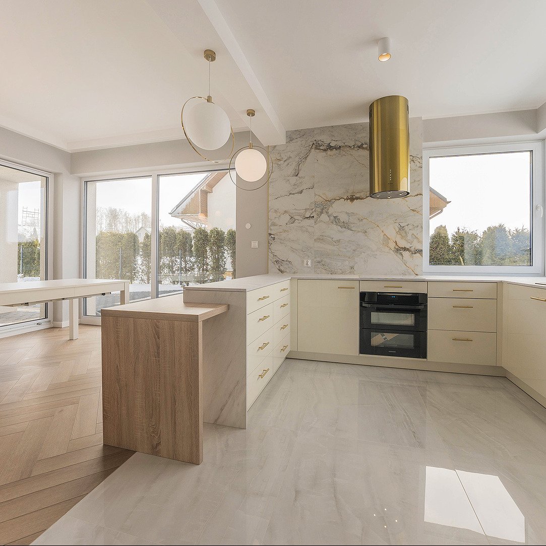 Jasna kuchnia w stylu nowoczesnym z wyspą, marmurowym panelem i złotym okapem. Duże okna wpuszczają naturalne światło. Widoczne lampy wiszące.
