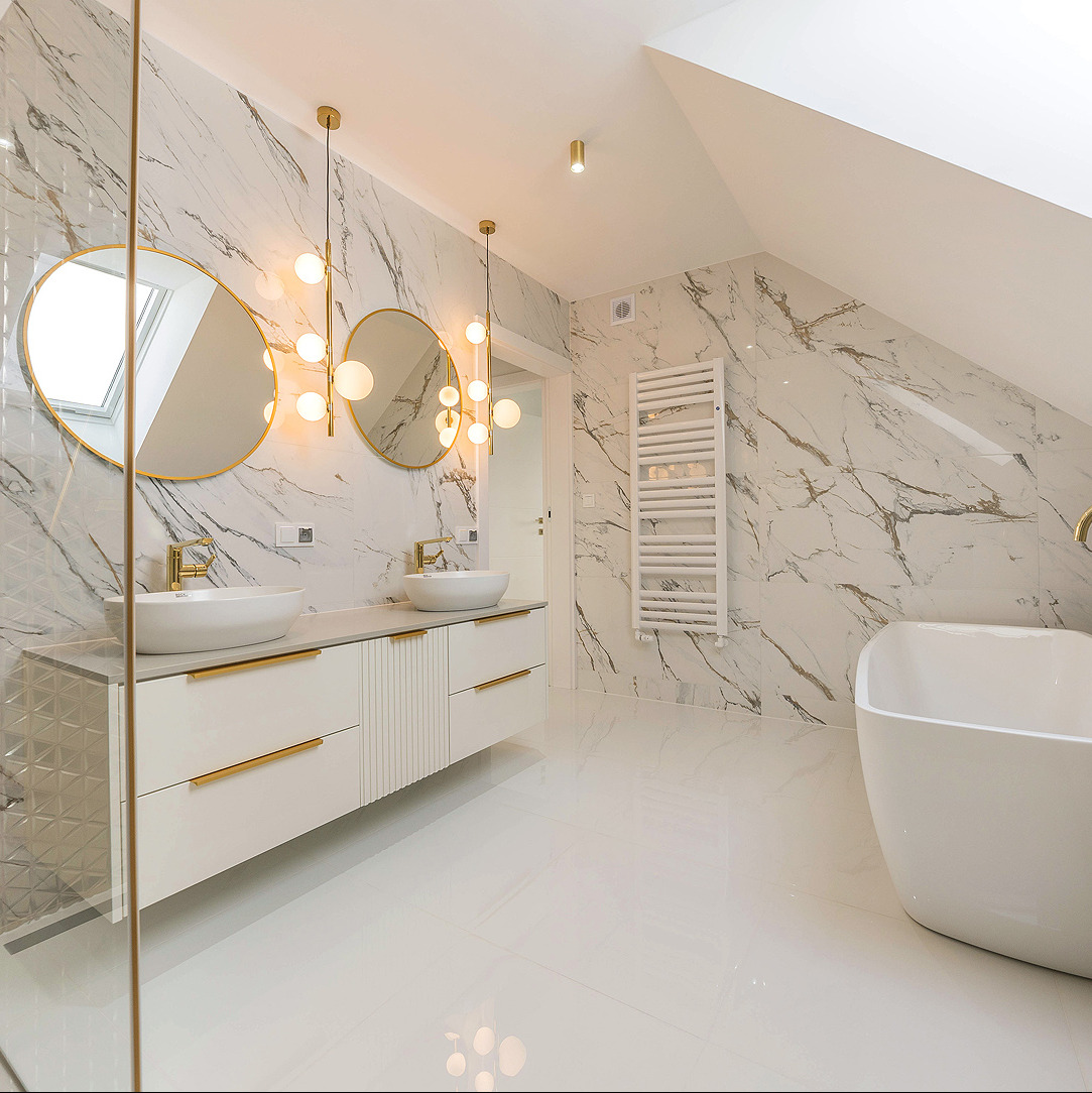Jasna łazienka na poddaszu z marmurowymi płytkami, podwójną umywalką z złotymi akcentami i wolnostojącą wanną. Nowoczesne oświetlenie kuliste.