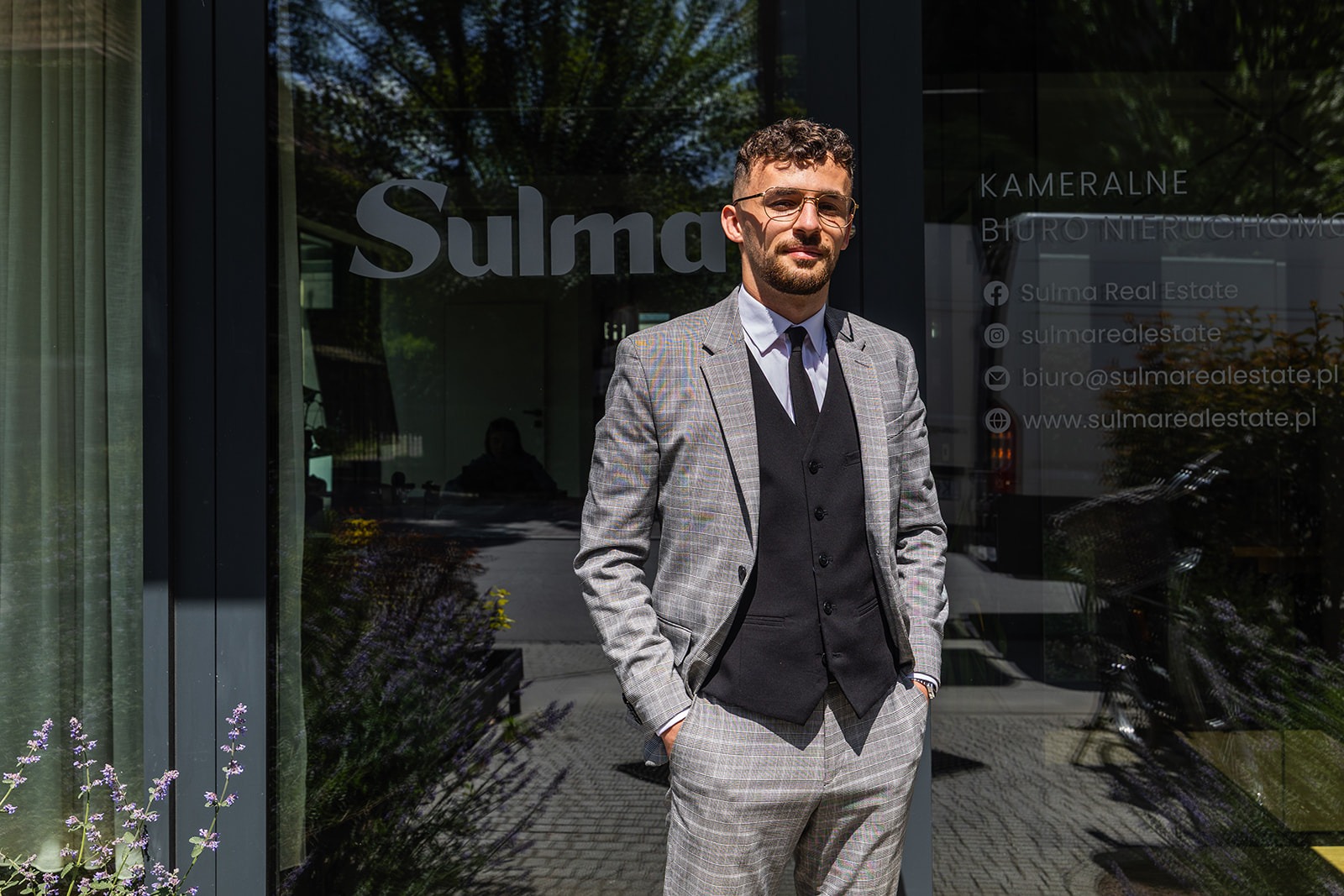 Młody mężczyzna w garniturze w kratę stoi przed biurem nieruchomości Sulma, z widocznym logo i danymi kontaktowymi na szklanej fasadzie.